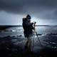 man-in-coat-and-hat-setting-up-tripod-on-beach-under-clouds